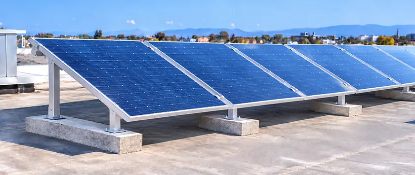 Rows of PV modules on adjustable tilt legs on a flat commercial roof, with taller rear legs showing adjustment holes and ballast blocks along the base rails.