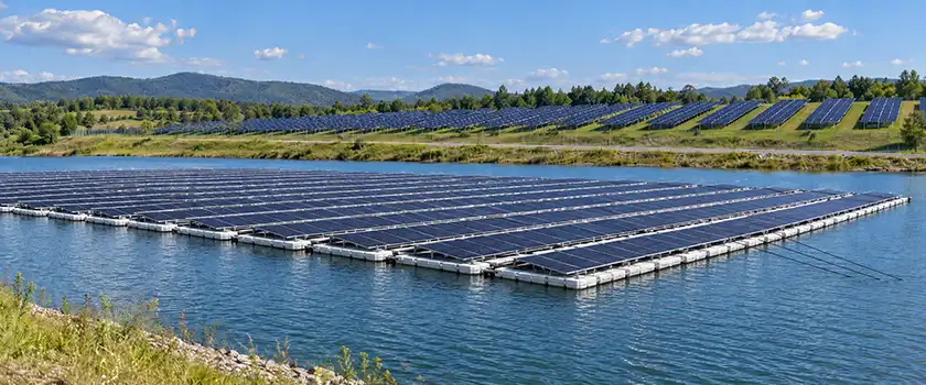 Floating solar array on HDPE floats on a reservoir, with fixed-tilt ground-mounted solar rows on the grassy shore behind it.